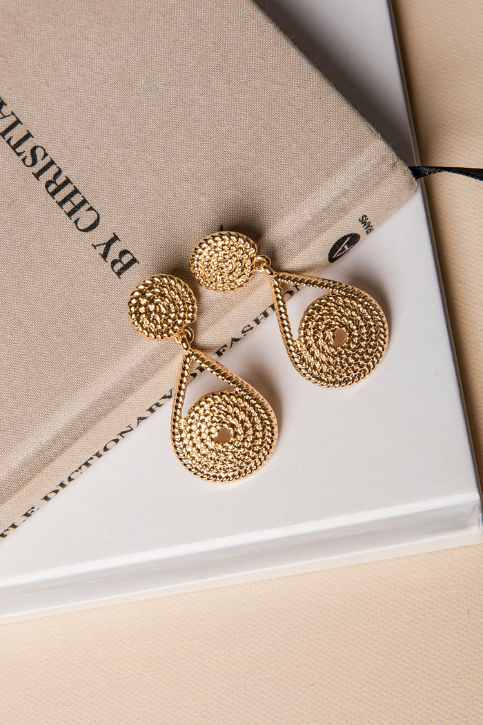 Gold earrings on a beige surface with 'By Christin' branding.