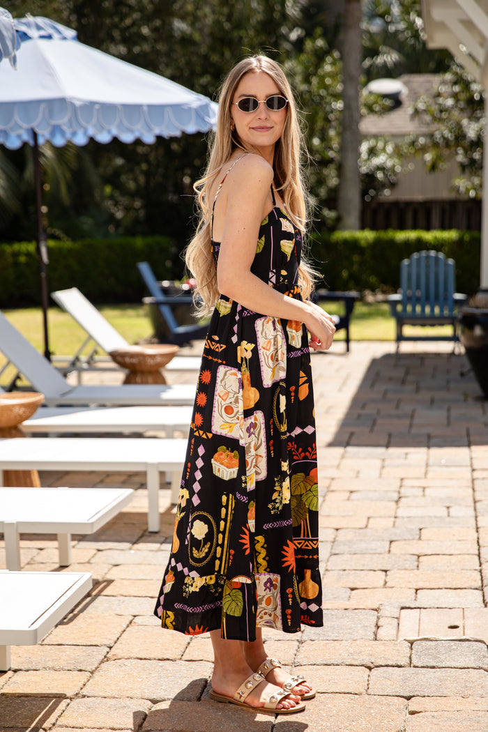 Woman in a patterned dress standing on a patio with lounge chairs and an umbrella in the background.