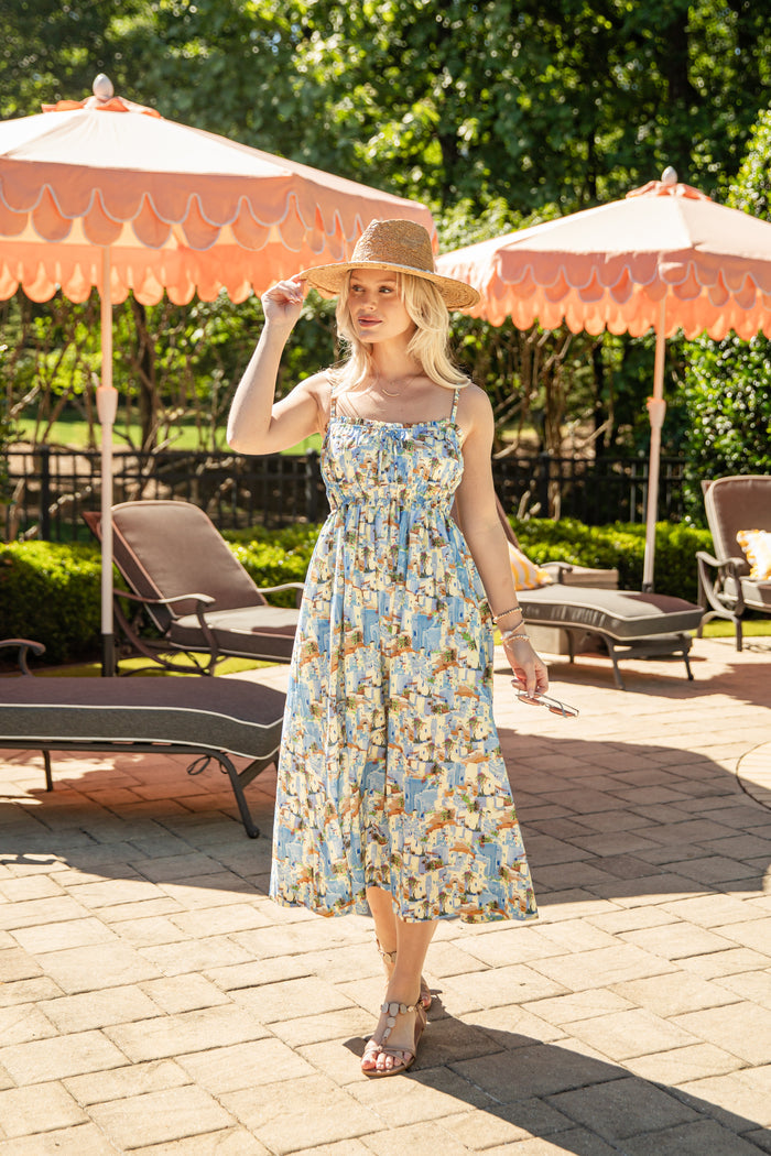 Woman in a floral dress standing outdoors with pink umbrellas and lounge chairs in the background.