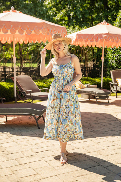Woman in a floral dress and hat standing on a sunlit patio with orange umbrellas and lounge chairs.
