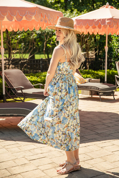 Woman in a floral dress standing under a patio umbrella with lounge chairs and greenery in the background.