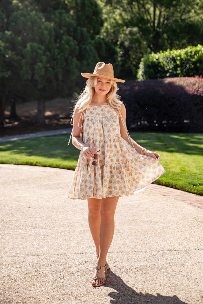 Woman in a patterned dress and hat standing on a paved path with greenery in the background