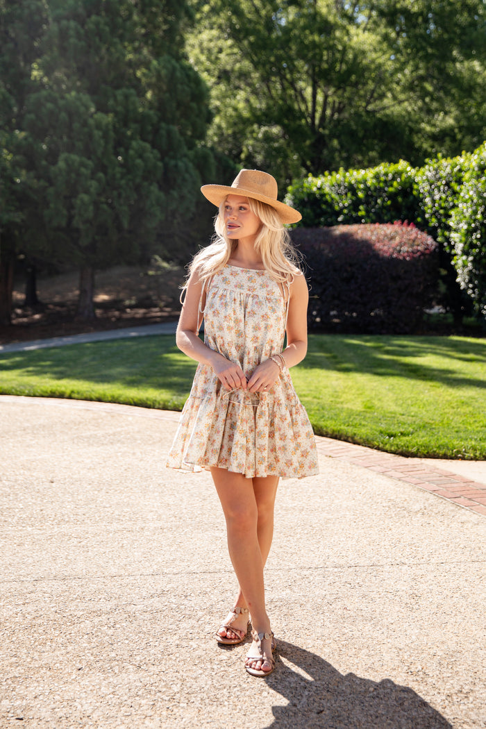 Woman in a floral dress and hat standing on a driveway with greenery in the background