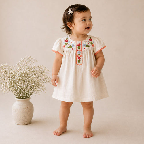 A toddler girl wearing a short-sleeve cream-colored dress with colorful floral embroidery on the chest and orange trim, standing barefoot next to a small vase of baby's breath against a warm beige background.