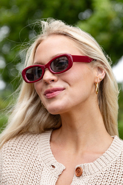 Woman wearing red sunglasses and a beige sweater outdoors with greenery in the background