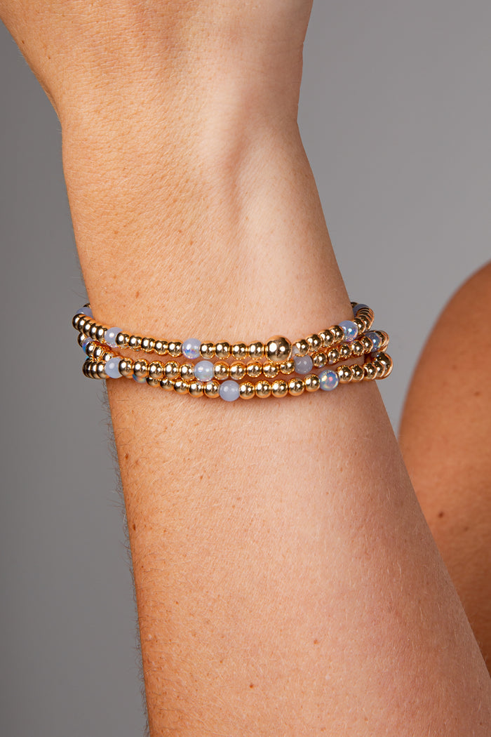 A close-up of a person's wrist wearing a stack of three beaded bracelets. The bracelets feature metallic gold-toned beads with small light blue-gray round accents.