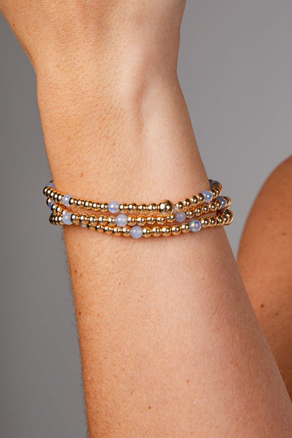 A close-up of a person's wrist wearing a stack of three beaded bracelets. The bracelets feature metallic gold-toned beads with small light blue-gray round accents.