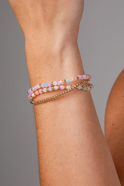 A close-up of a person's wrist wearing three stacked bracelets: two beaded bracelets in pastel pink, peach, and light blue, and one thin gold chain bracelet.