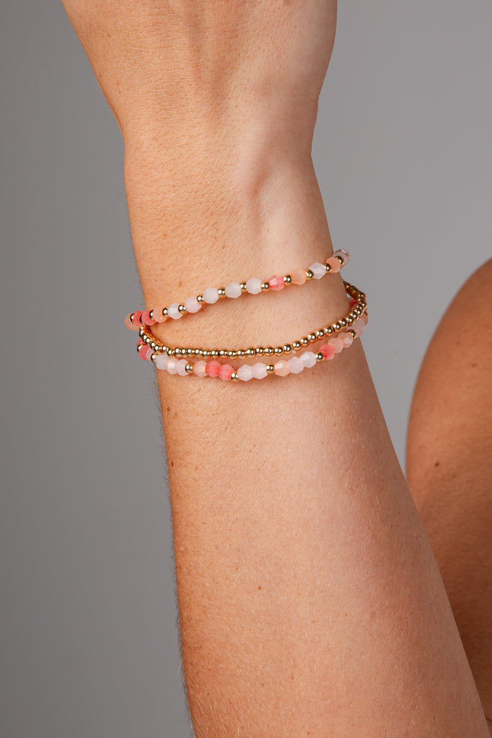 A close-up of a person's wrist wearing three beaded bracelets: two with pink and white beads separated by small gold spacer beads, and one with a continuous gold chain.