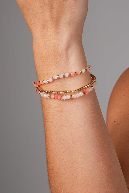 A close-up of a person's wrist wearing three beaded bracelets: two with pink and white beads separated by small gold spacer beads, and one with a continuous gold chain.