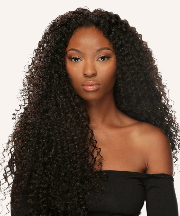 A woman with long, voluminous curly black hair in an off-the-shoulder black top stands before a light background, looking directly at the camera with a neutral expression and effortless poise.